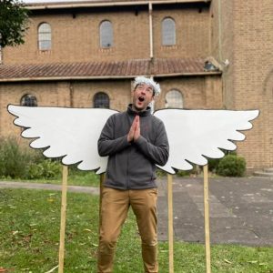 James Faragher posing with hands clasped as though praying, with a joking expression on his face, in front of a pair of angel wings made of wood