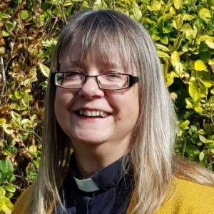 A blonde lady with glasses, wearing a clerical collar and a mustard jumper smiles into the camera