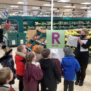 A group of children standing in Tescos with a woman in a facemask demonstrating reusable veggie bags