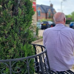 A man in a shirt sits on a metal bench which surrounds a tree in a circular shape. He looks out across a carpark