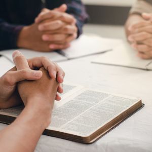 Three pairs of hands clasped in prayer over bibles on a table