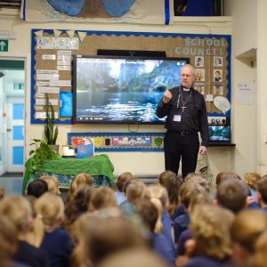 Archbishop Justin stands with one hand lifted and clenched and a bemused expression, in front of a screen where a scene with water is showing.
