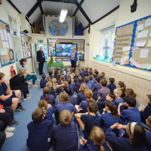 A view from the back of a classroom. The Archbishop and headteacher Kathryn Wilson stand at the front of the room addressing children who are wearing blue t shirts and jumpers.